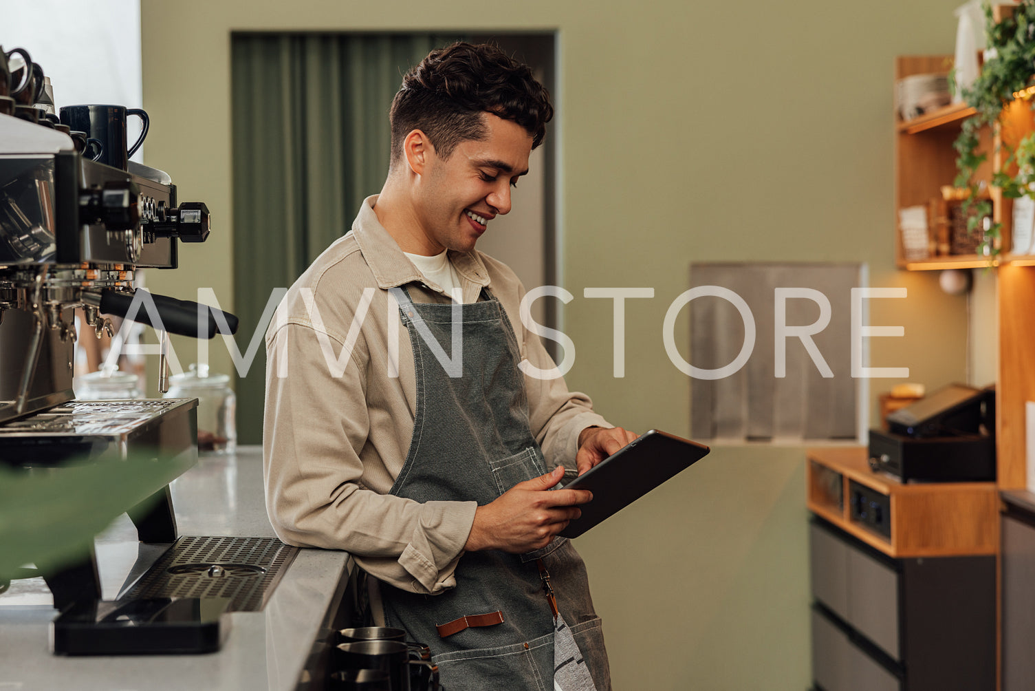 Side view of a smiling barista in an apron typing on a digital tablet leaning counter in a coffee shop