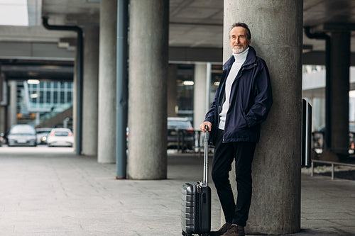 Stylish mature tourist standing outdoors. Man with a suitcase leaning on the wall looking away.