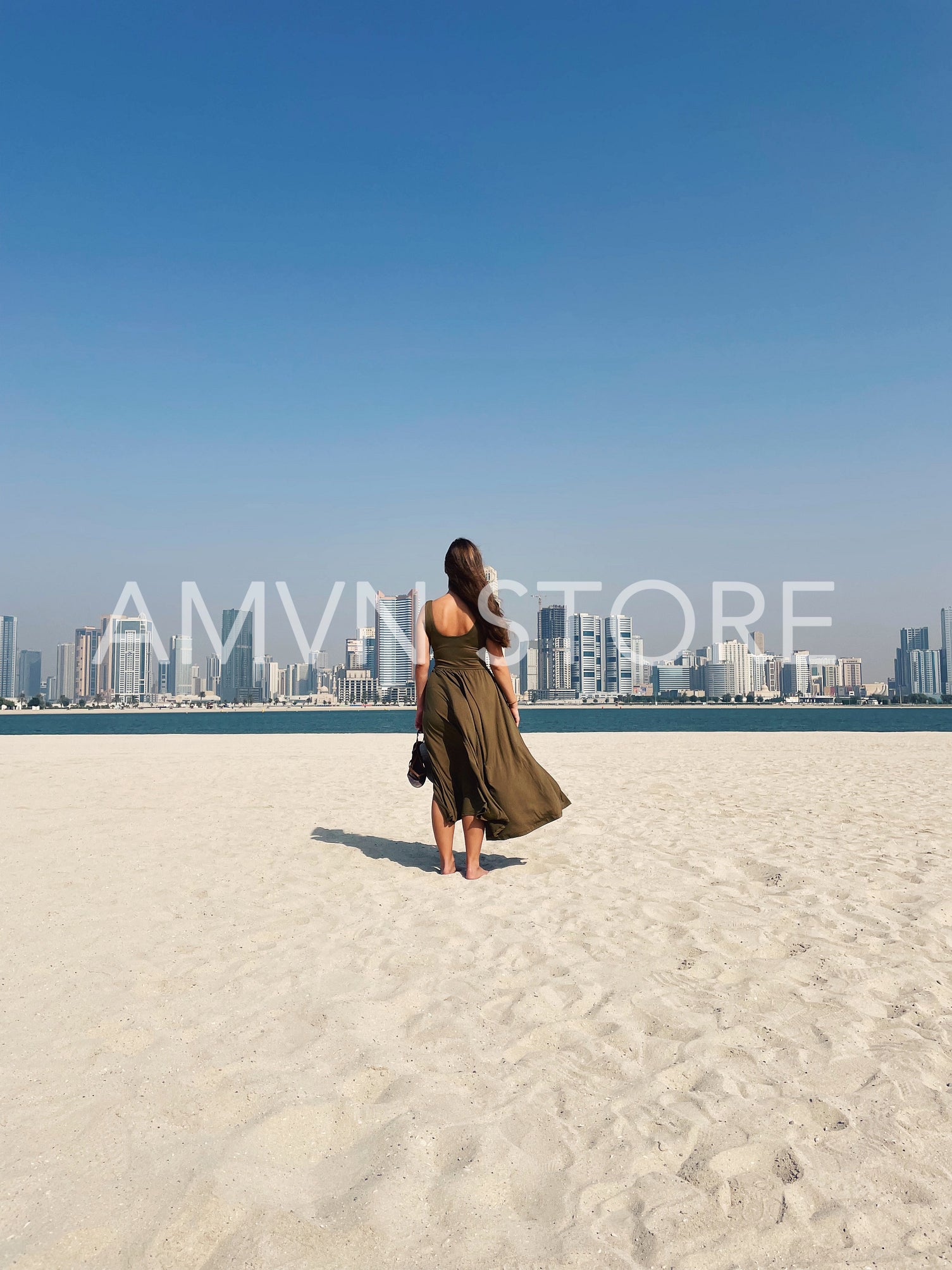 Back view of young female in dress standing on a beach against a cityscape with skyscrapers