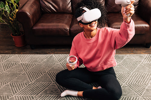 Smiling woman in living room using VR set. Young female in comfy clothes playing video games while sitting on the floor.