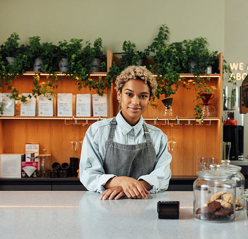 Young woman barista in an apron looking at the camera while standing at a counter in a small cafe