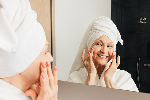 Smiling aged woman with a towel on a head applying facial cream on her cheeks in bathroom