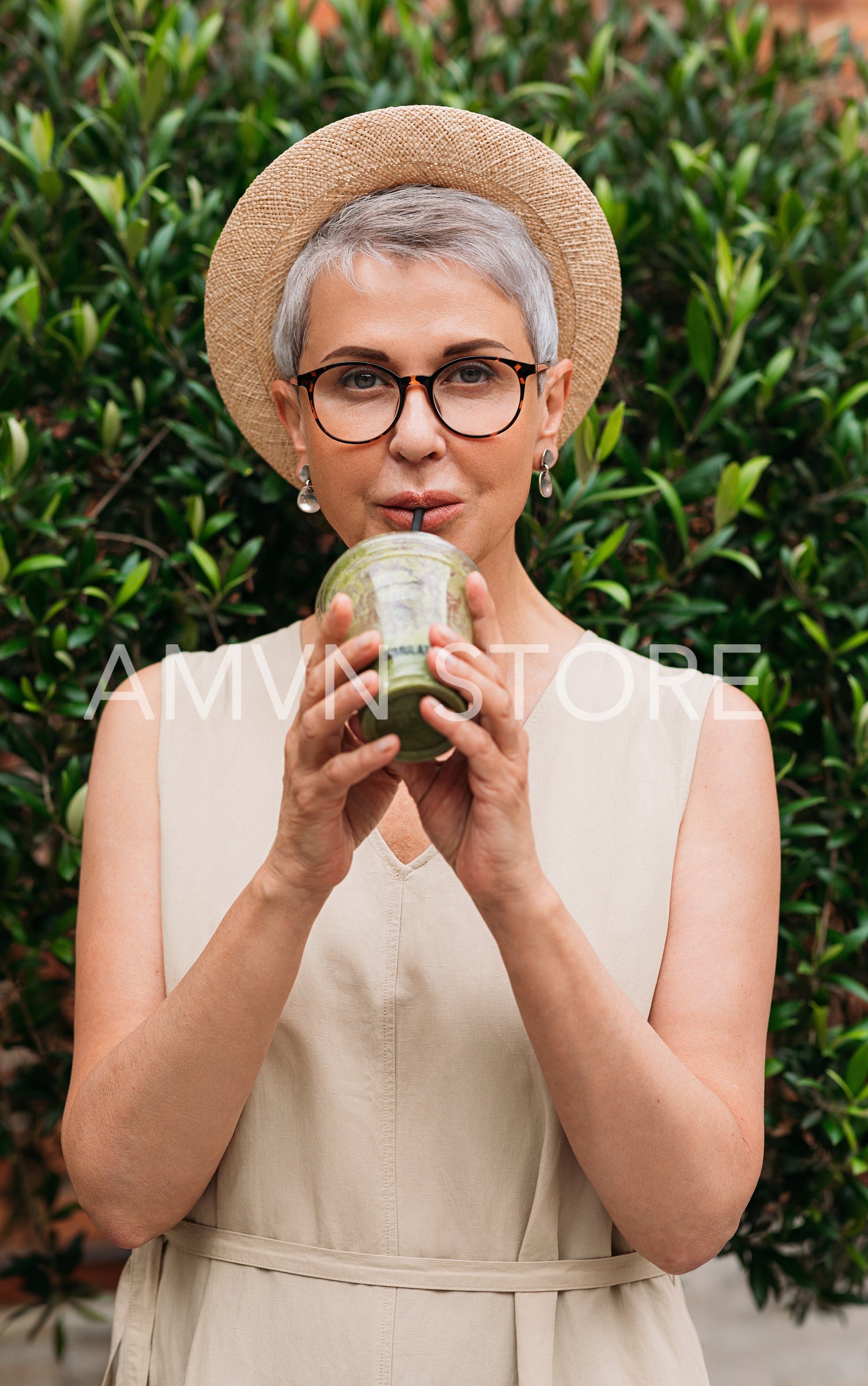 Stylish mature woman in a straw hat drinking a green smoothie in the park