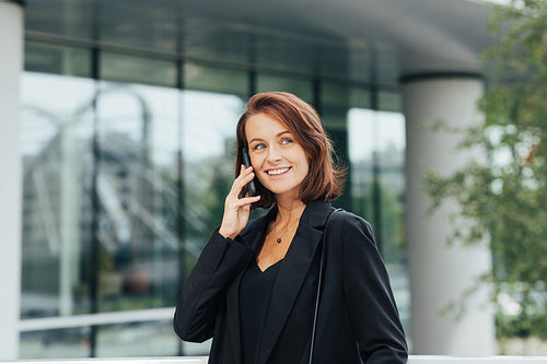 Cheerful female entrepreneur talking on a mobile phone. Woman in black formal wear is looking away while having a conversation on a cell phone.