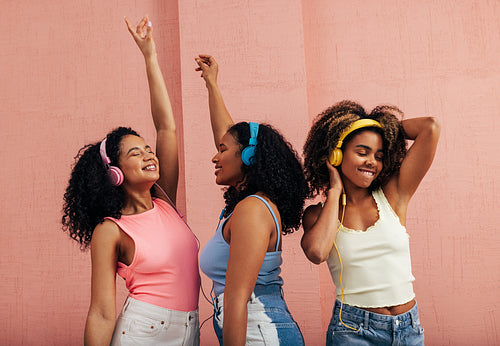 Three girlfriends with different body types dancing together. Smiling females with headphones enjoying music at pink wall.