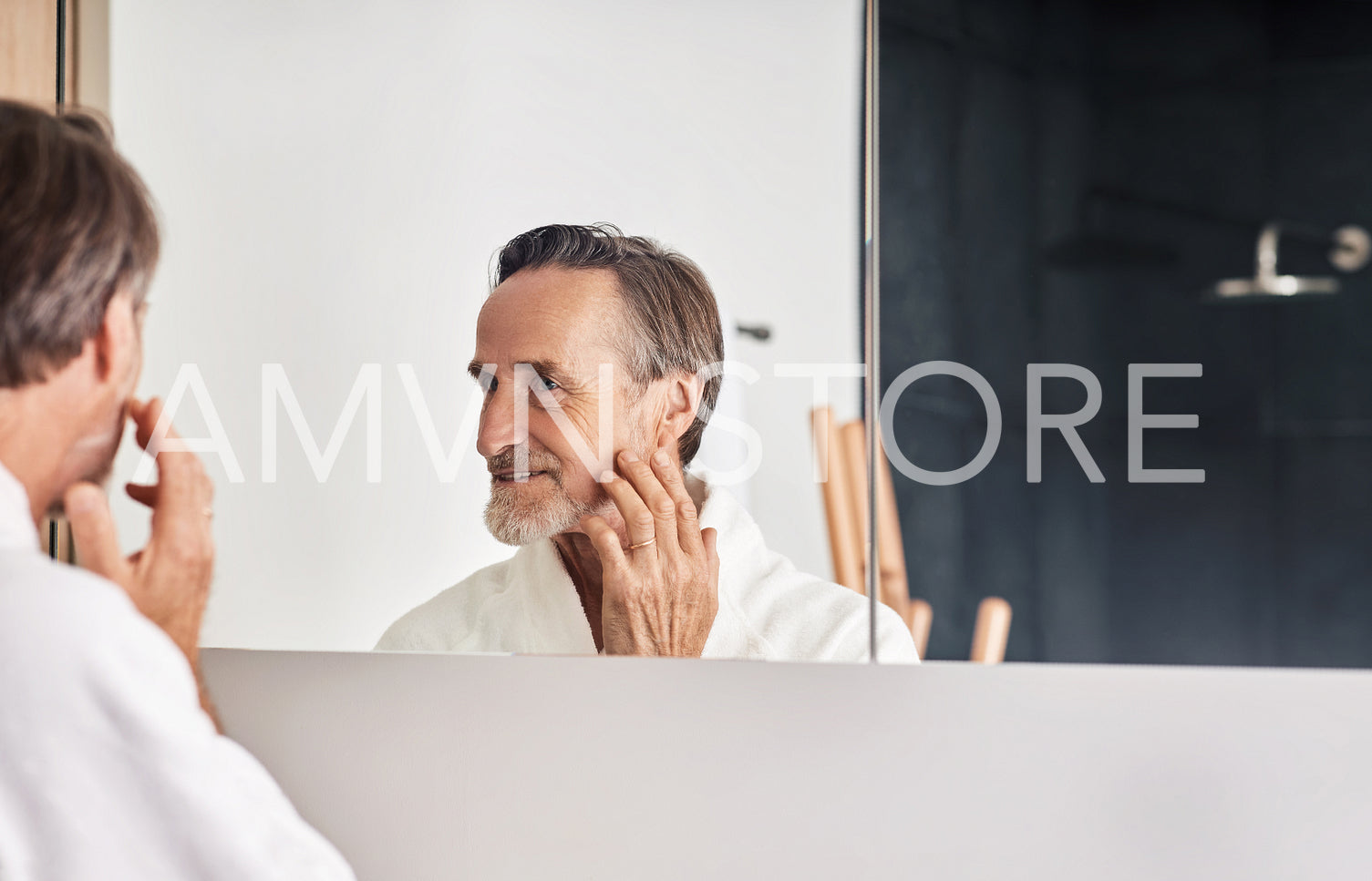 Smiling senior man looking at his reflection in the mirror and touching face. Mature male in a bathroom in the morning.	