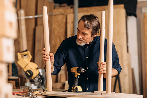 Senior man observing table legs in the workshop. Carpenter working on furniture.