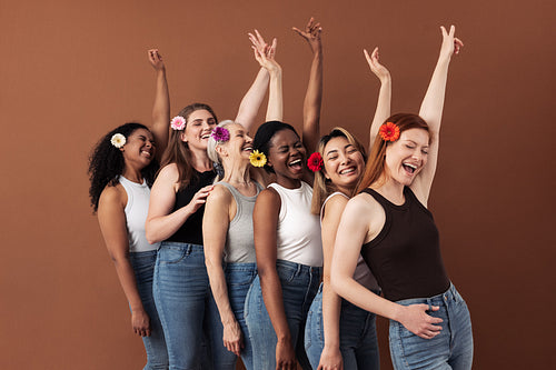 Six laughing women with flowers in hair. Multi-ethnic group of females raises hands up over brown background.