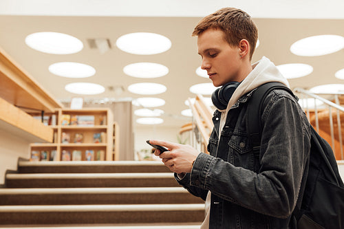 Young male with backpack and smartphone standing in library