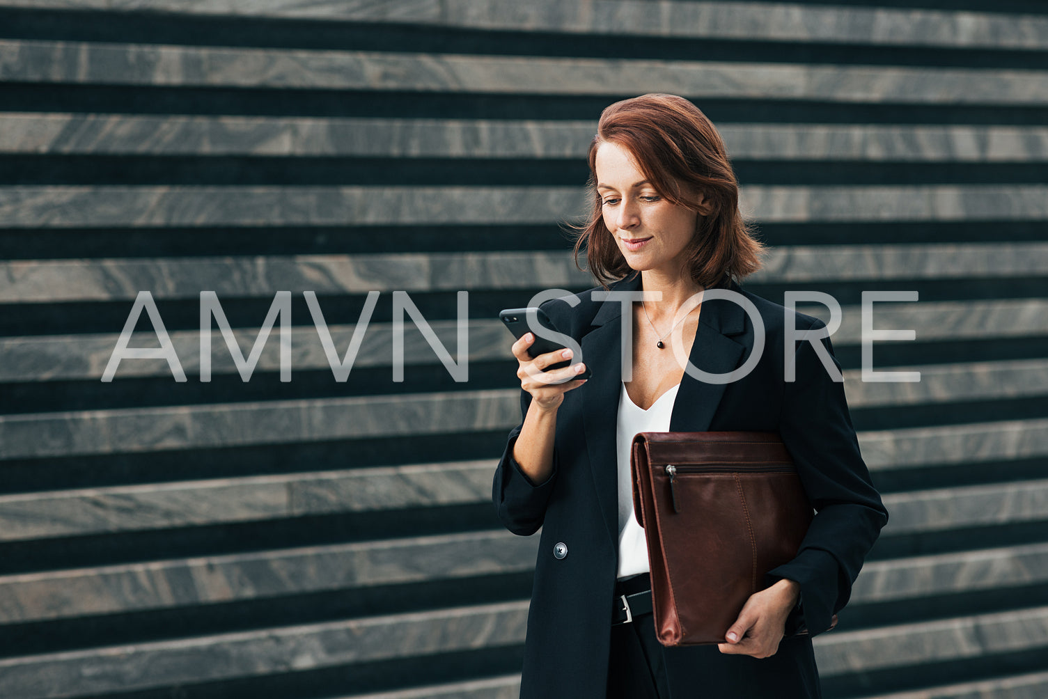 Smiling businesswoman holding a smartphone and leather folder standing outdoors. Middle-aged female with ginger hair typing on smartphone.