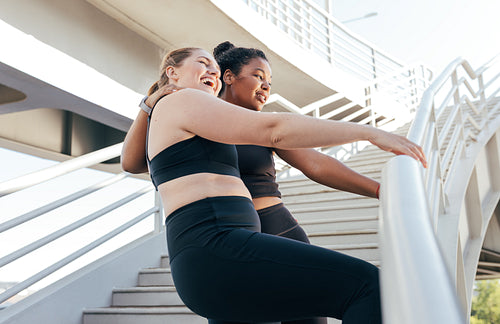 Two sportswomen having fun while standing at railing on the bridge after an intense workout. Female friends in sportswear enjoying a good mood after exercises.