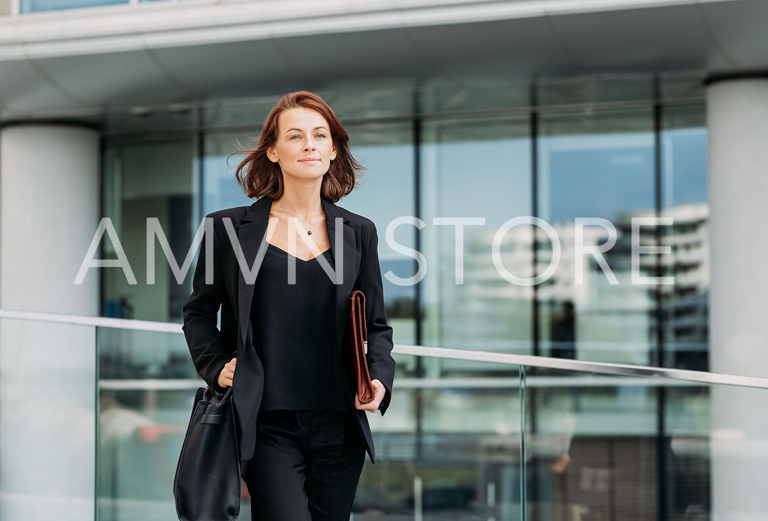 Confident female with a bag and leather clothes walking outdoors against a glass building. Cheerful corporate person walking outdoors.