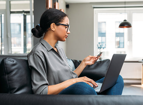 Woman using credit card for paying online, sitting on a sofa in the living room