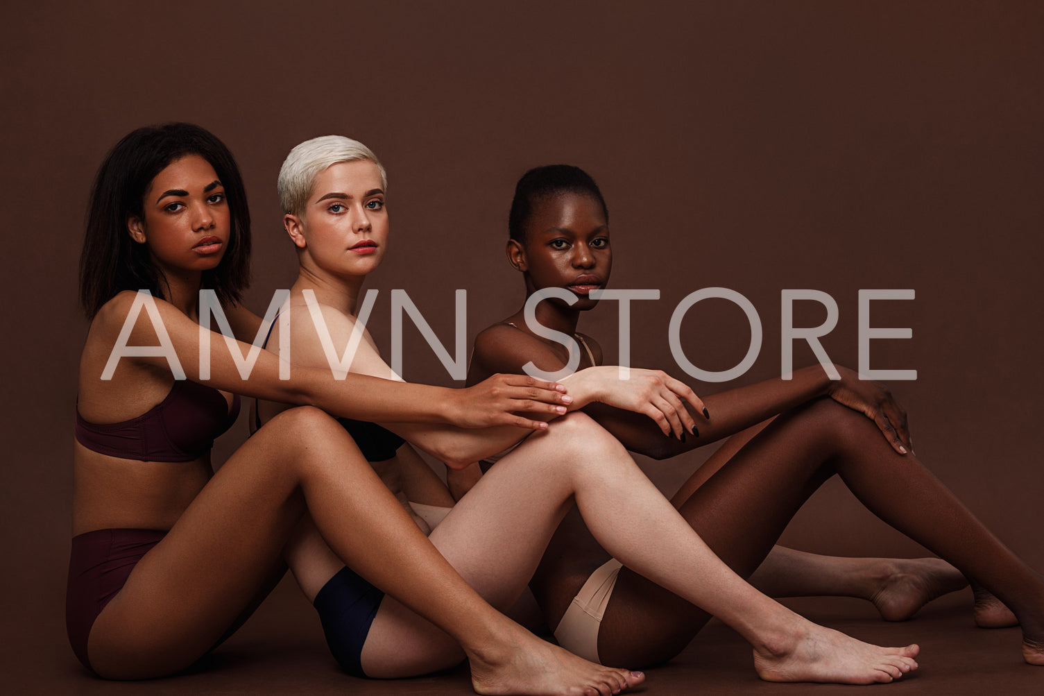 Side view of three diverse women sitting together in lingerie on brown background and looking at camera