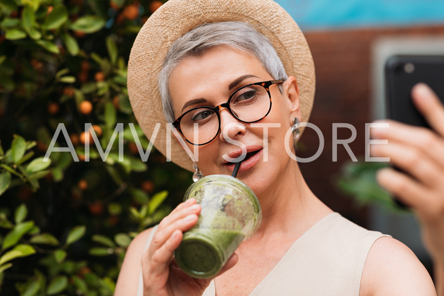 Senior woman taking a selfie outdoors while drinking a green smoothie. Portrait of a mature female in a small straw hat taking a selfie in the park.