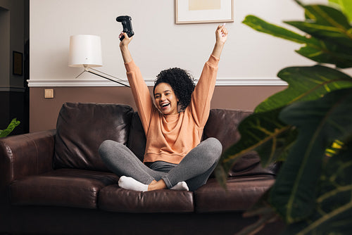 Happy mixed race woman sitting on a sofa in living room with raise hands up and holding a joystick