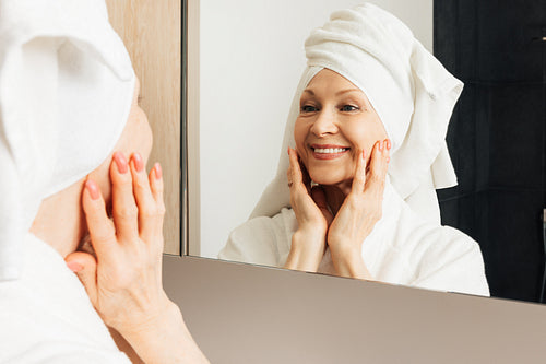 Portrait of a senior female with a towel on her head. Aged woman touching face in front on a mirror.