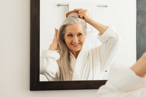 Smiling senior woman combing her grey hair in front of a bathroom mirror