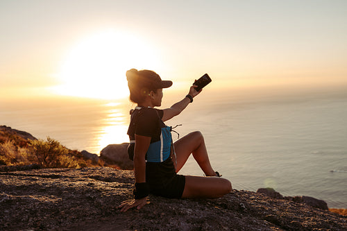 Young fit woman in sportswear taking selfie while sitting on a hill at sunset