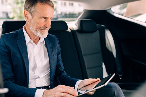 Side view of businessman sitting on a backseat of car and using digital tablet