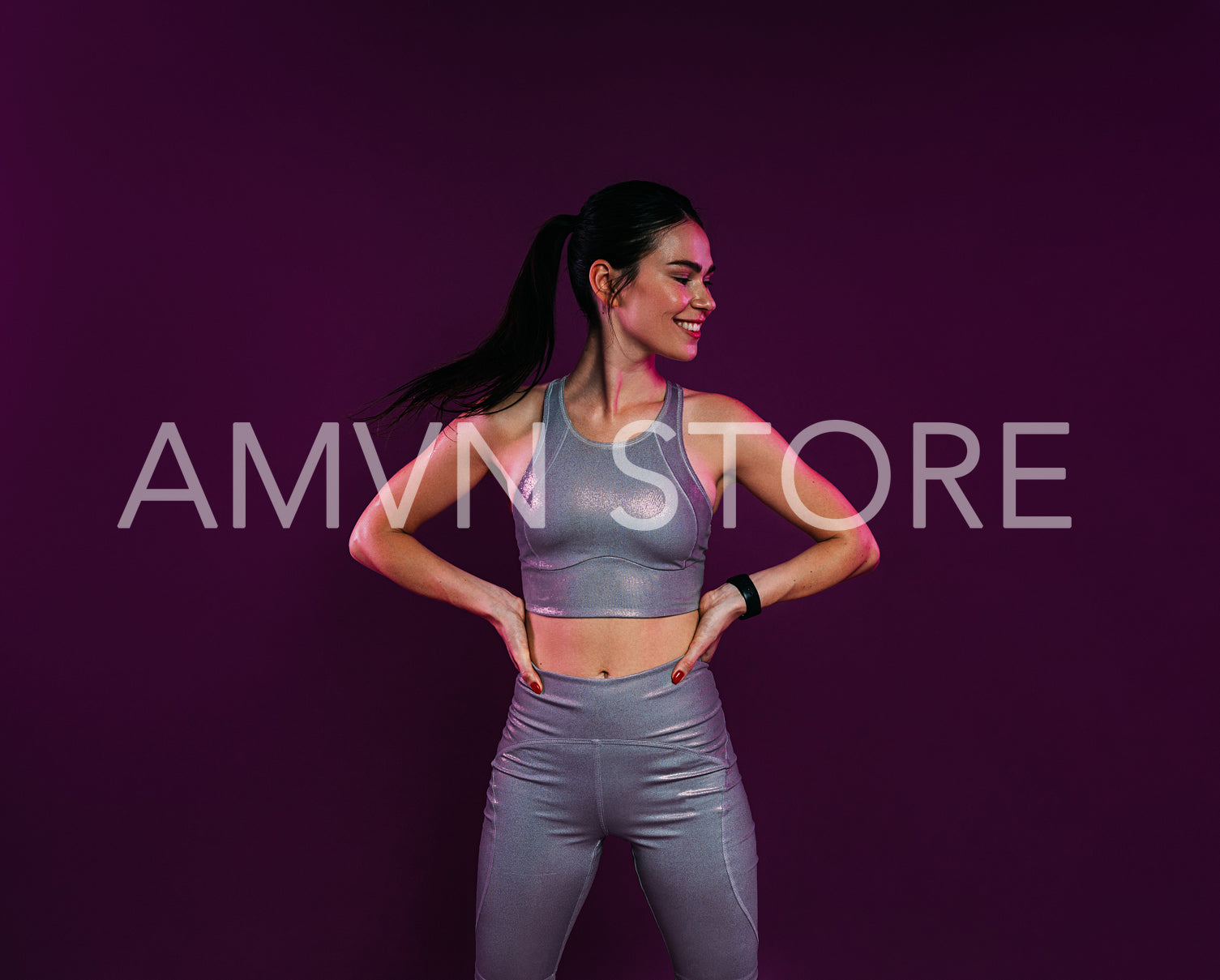 Young woman in silver fitness attire with hands on hips looking away at magenta background