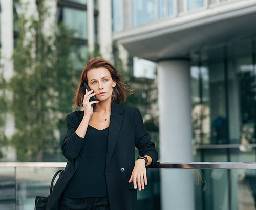 Confident middle-aged businesswoman with ginger hair leaning on railing talking on mobile phone outdoors