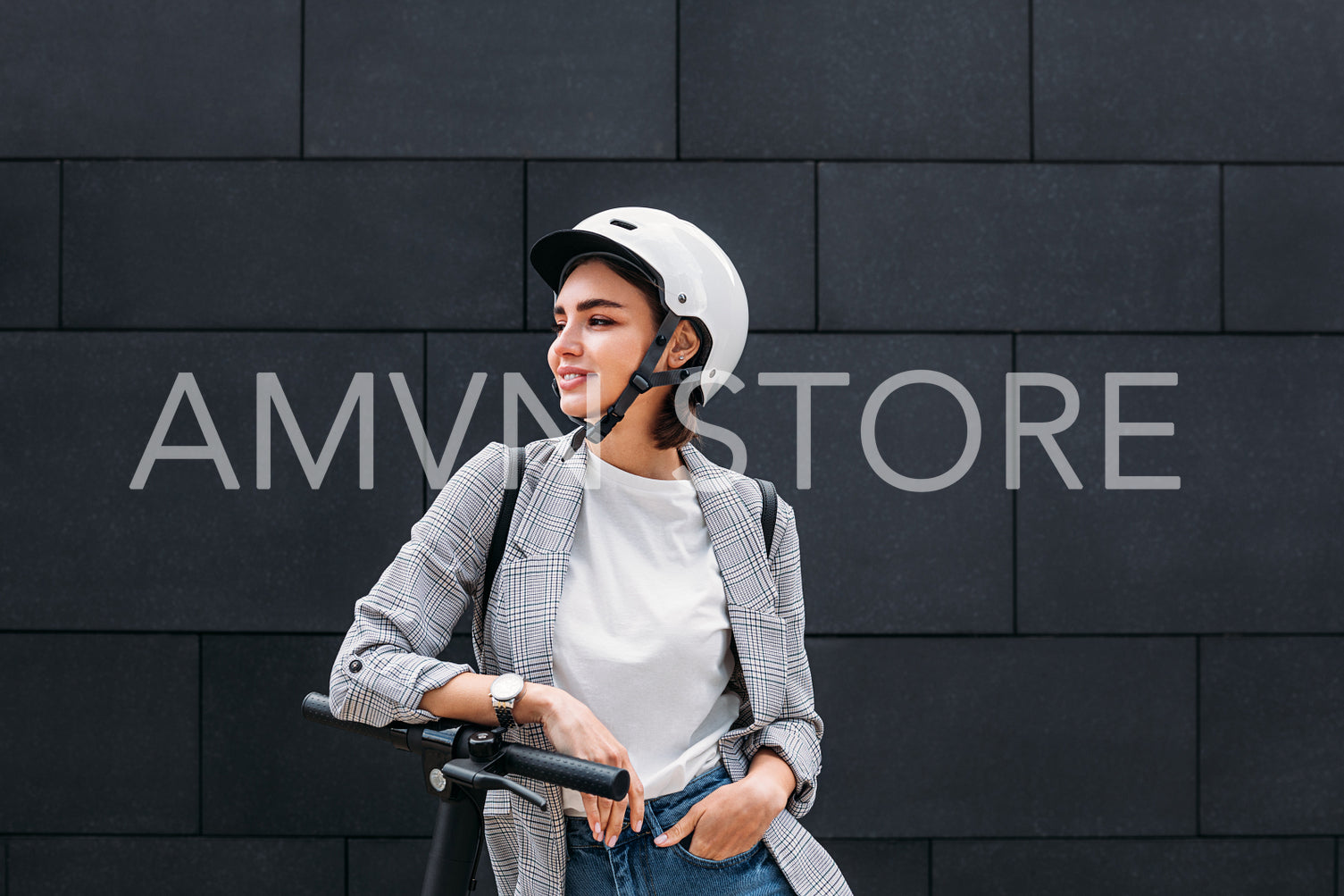 Young woman wearing a scooter helmet standing at a black wall. Smiling female in casual clothes leaning electric scooter looking away.