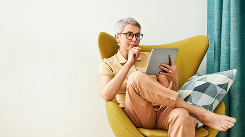 Stylish mature woman in eyeglasses thoughtfully looking at a digital tablet while sitting at home in an armchair