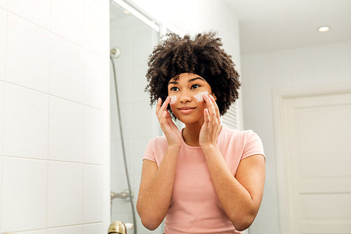 Mixed race woman in bathroom looking into the mirror and applying moisturizer on her face