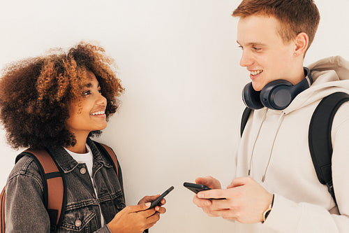 Two classmates standing at wall and looking to each other holding smartphones