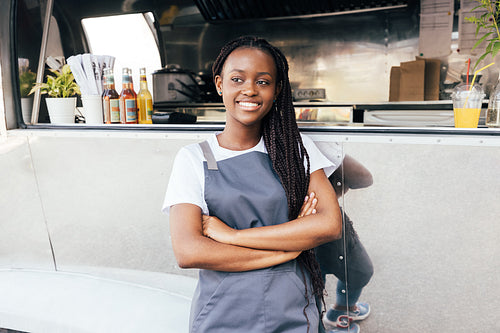 Smiling female entrepreneur wearing apron standing at a food truck. Saleswoman with arms crossed looking away.