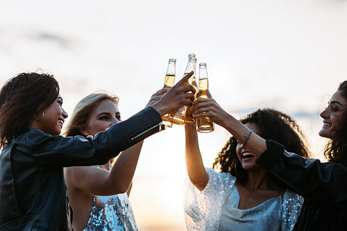 Multi-ethnic friends toasting beers at sunset. Women celebrating with beers in the evening.