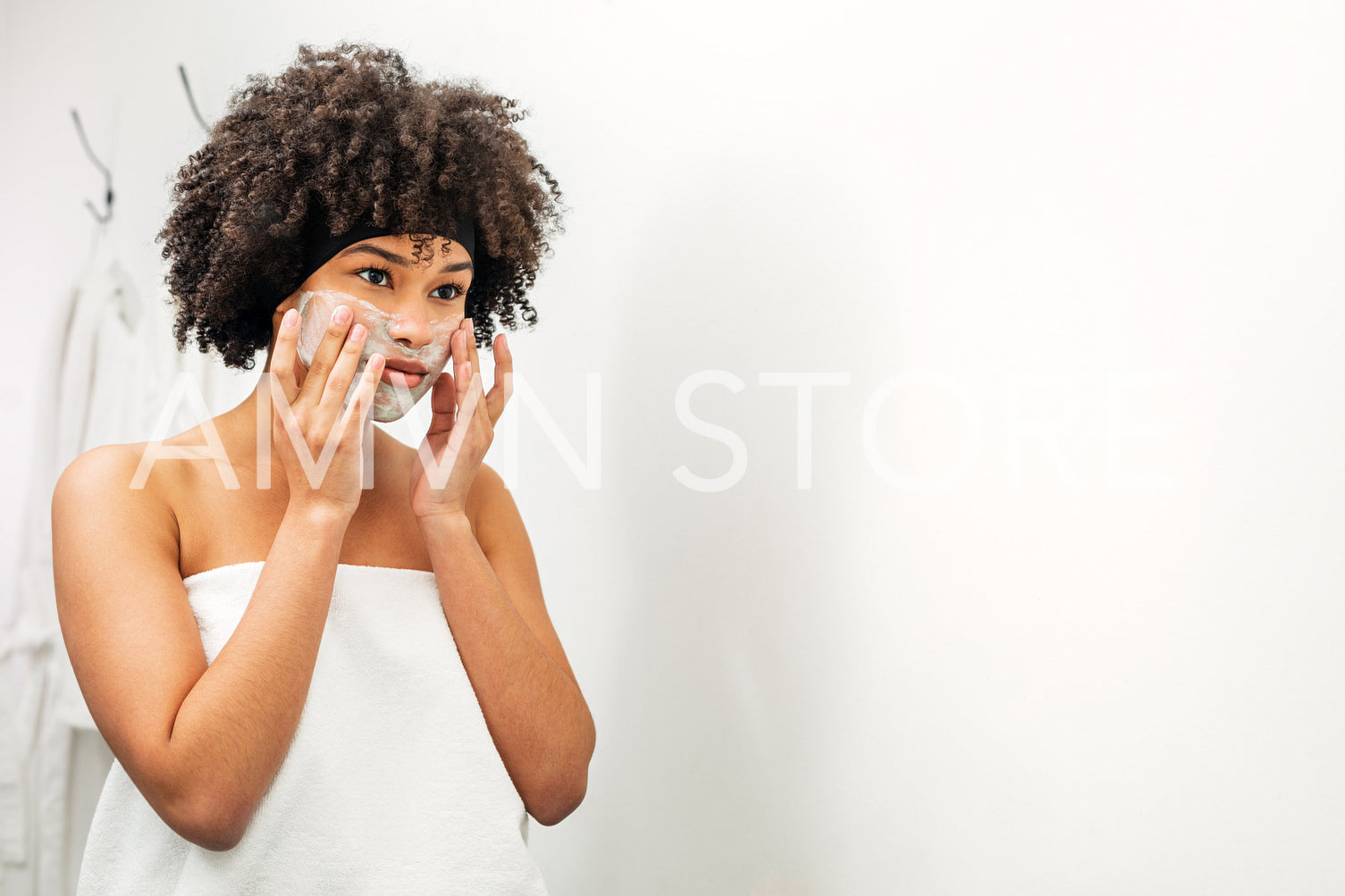 Young woman wrapped in white towel applying natural mask on her face	