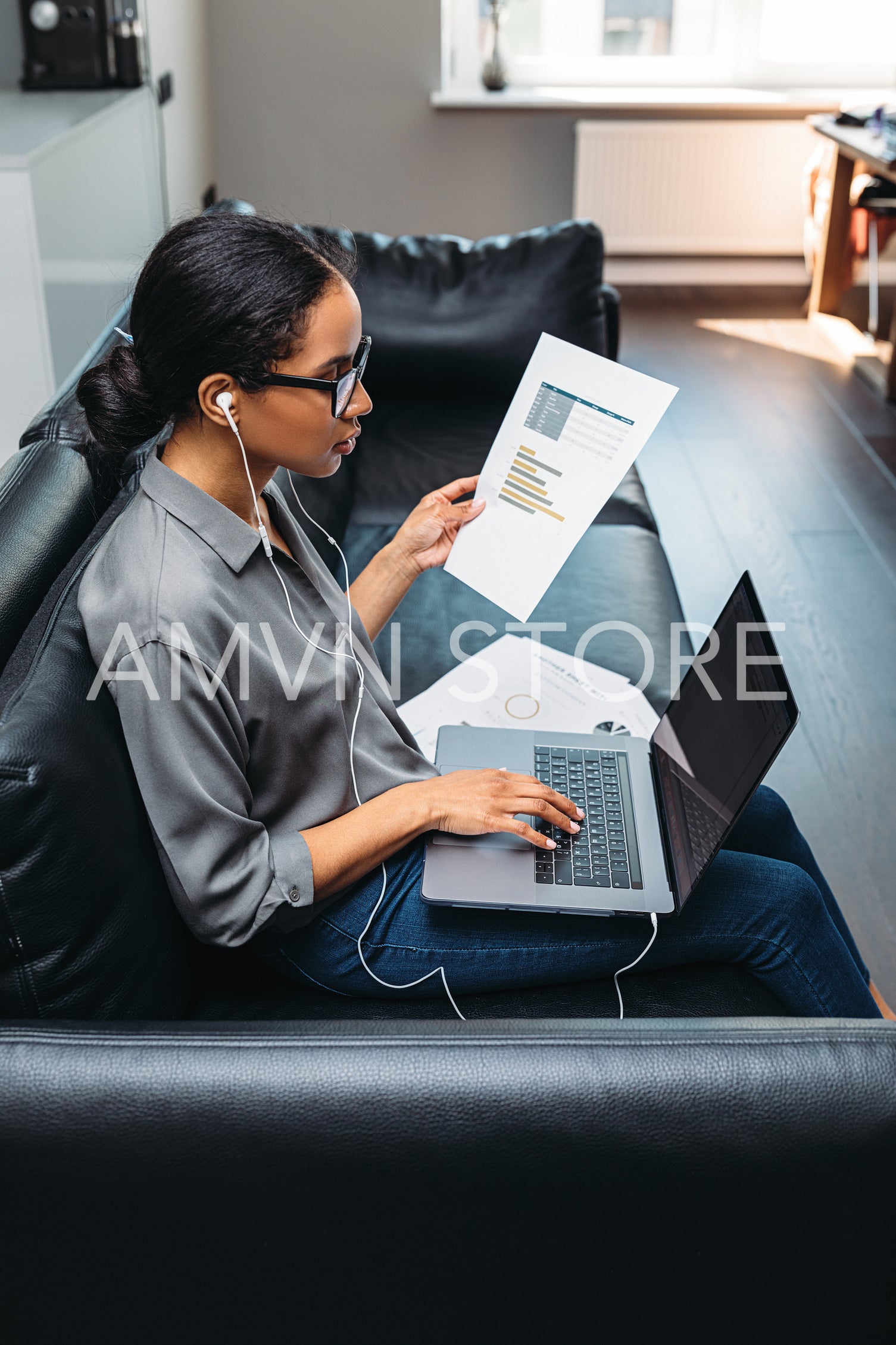 Female entrepreneur using laptop sitting on a sofa wearing earphones. Side view of a young businesswoman working from home.	