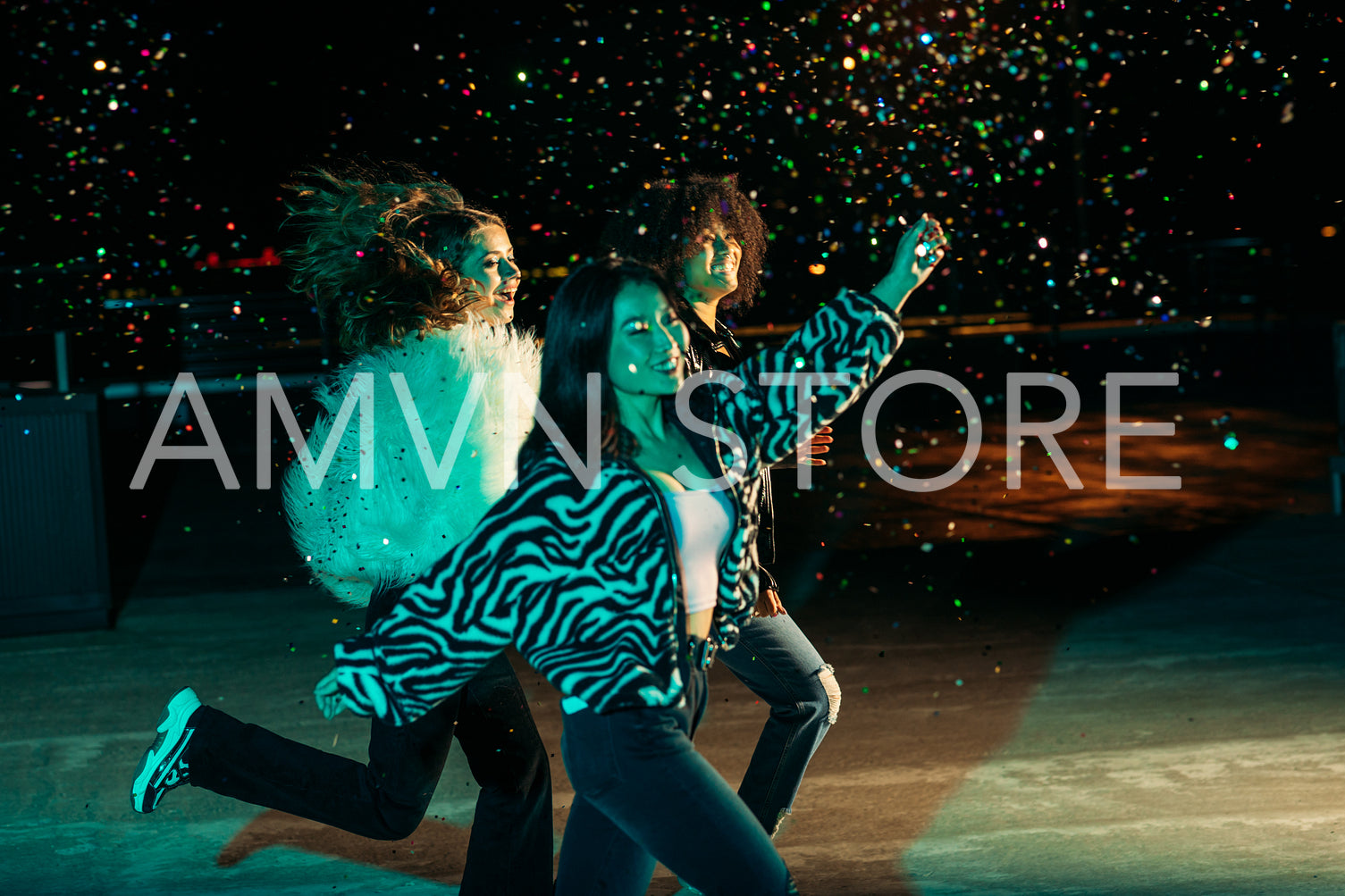 Happy female friends running together on the roof and throwing confetti at night. Three stylish girls celebrating outdoors.	