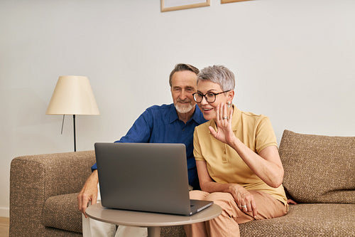 Cheerful senior couple using a laptop for video calling from the living room. Woman and man sitting