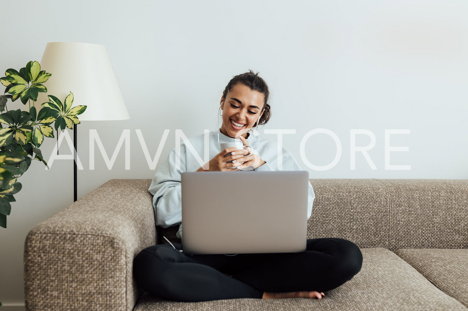 Young smiling woman holding a cup while sitting on a sofa with laptop on legs	