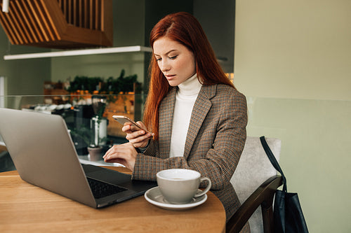 Woman with ginger hair holding a smartphone sitting at a cafe. Businesswoman working from the coffee shop.