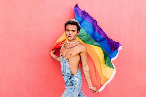 Portrait of a handsome young man holding pride movement LGBT rainbow flag against a pink wall. Man with a pride flag looking at camera outdoors.