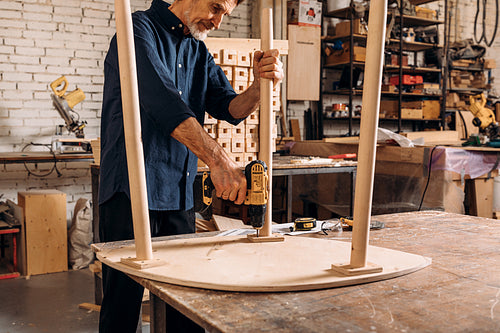 Senior male carpenter drilling a hole in wooden table standing at workbench