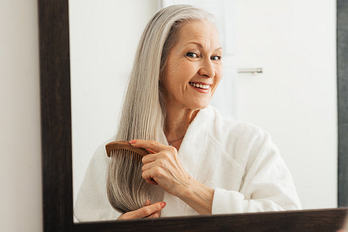 Woman combing her hair in bathroom. Senior female using a wood comb.