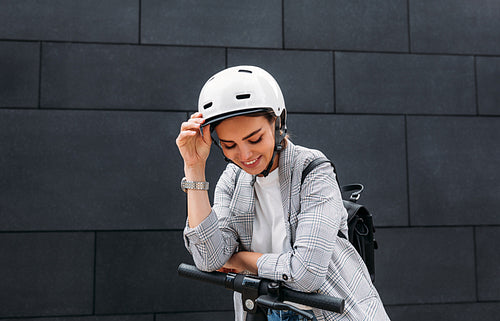 Happy businesswoman in cycling helmet leaning on handlebar of electric push scooter looking down