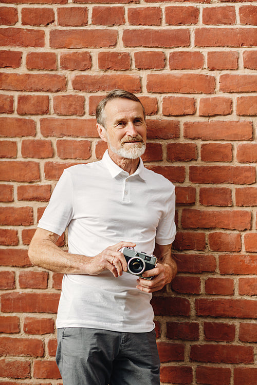 Senior man standing with a film camera outdoors at a brick wall