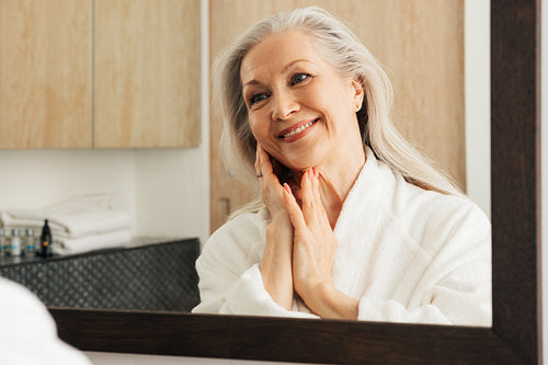 Cheerful woman with grey hair admires her reflection in a mirror