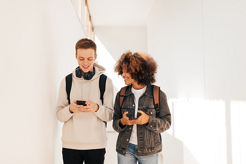 Two smiling students walking in corridor and chatting while holding smartphones