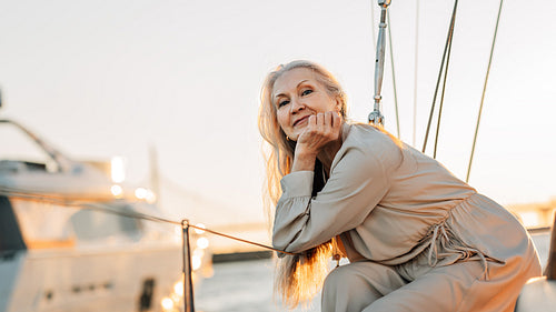 Portrait of a mature Caucasian woman with long grey hair looking straight to camera while sitting on a sailboat at sunset