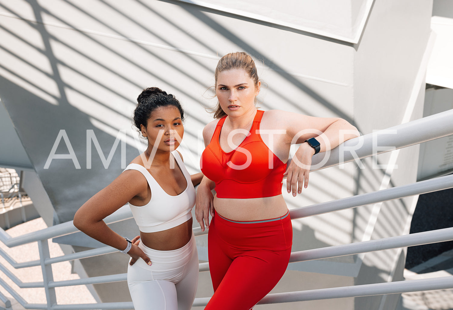 Portrait of two plus-size females in sportswear standing together. Two women in fitness attire standing on the stairs of a bridge and looking at camera.