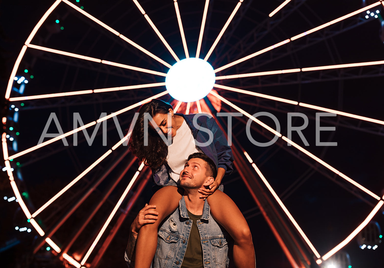 Smiling woman looking at her boyfriend while sitting on his shoulders at night in an amusement park