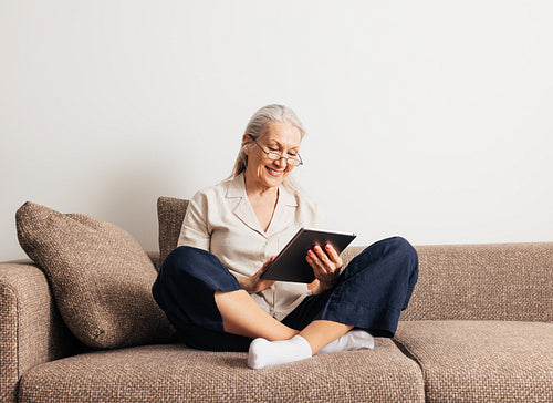 Cheerful aged female in glasses relaxing on a couch holds a digital tablet