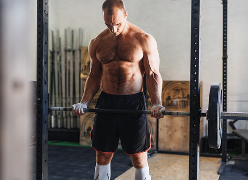 Muscular man exercising with barbell in gym, looking at his arms